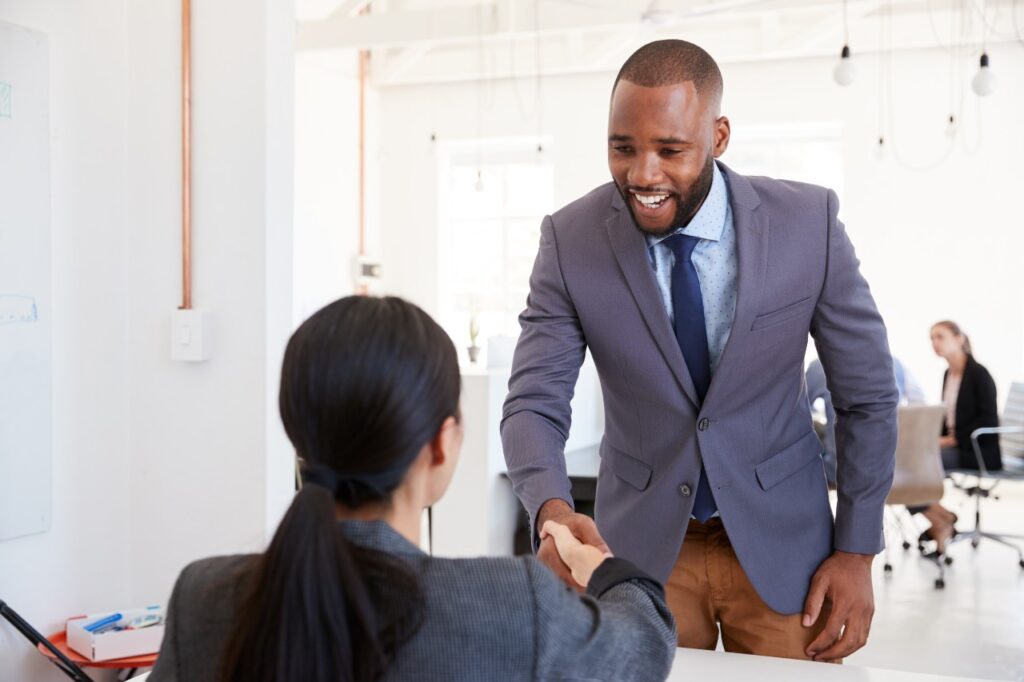 Black businessman and seated woman shaking hands in office hiring tech talent