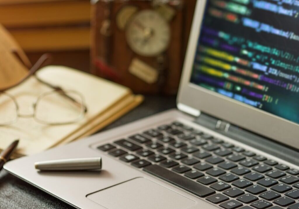 Laptop with programming code, book, pen and clock at office desk. Low depth of field.